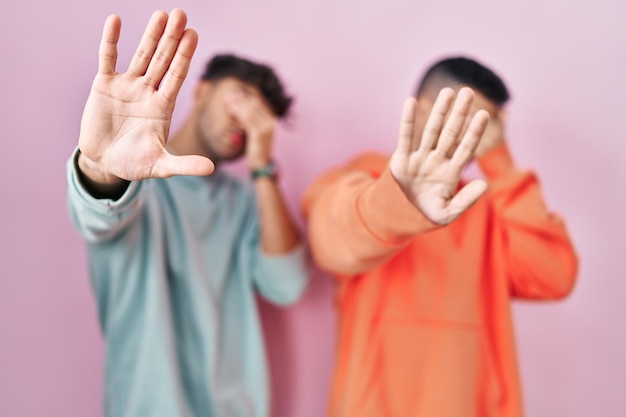 Young hispanic gay couple standing over pink background covering eyes with hands and doing stop gesture with sad and fear expression. embarrassed and negative concept.