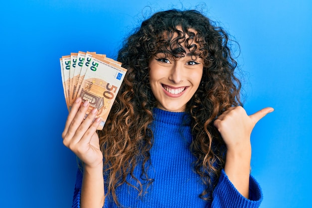 Giovane ragazza ispanica che tiene un mazzo di banconote da 50 euro che punta il pollice verso l'alto sorridendo felice con la bocca aperta