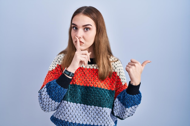 Young hispanic girl standing over blue background asking to be quiet with finger on lips pointing with hand to the side silence and secret concept