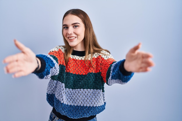 Young hispanic girl standing over blue background looking at the camera smiling with open arms for hug. cheerful expression embracing happiness.