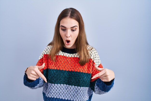 Young hispanic girl standing over blue background pointing down with fingers showing advertisement surprised face and open mouth