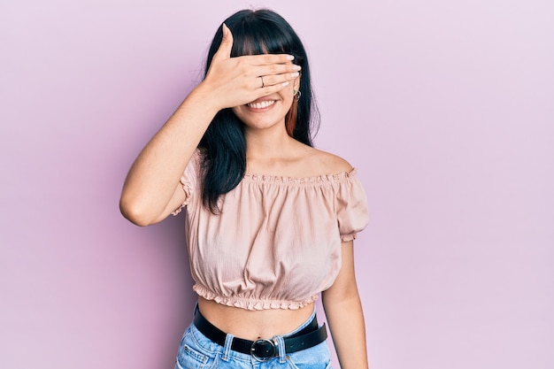 Young hispanic girl wearing casual clothes smiling and laughing with hand on face covering eyes for surprise blind concept