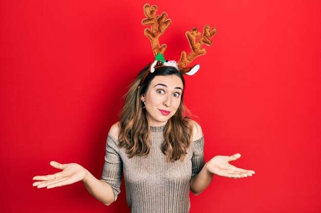 사슴 크리스마스 모자를 쓰고 팔과 손으로 혼란스러운 표정을 짓고 있는 젊은 히스패닉 소녀는 의심의 개념을 제기했습니다.