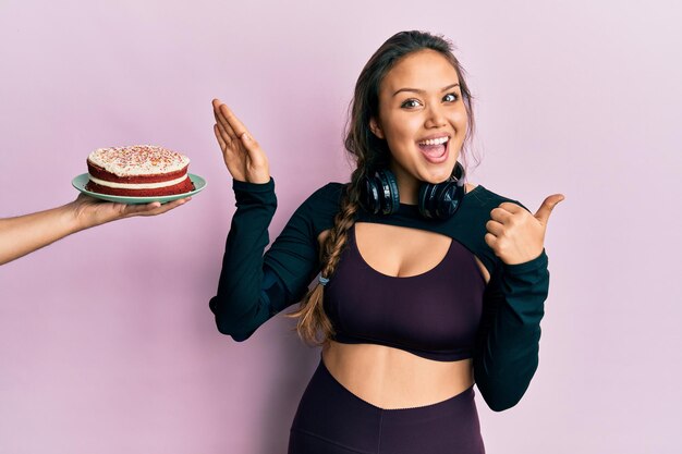 Young hispanic girl wearing sportswear rejecting cake pointing thumb up to the side smiling happy with open mouth