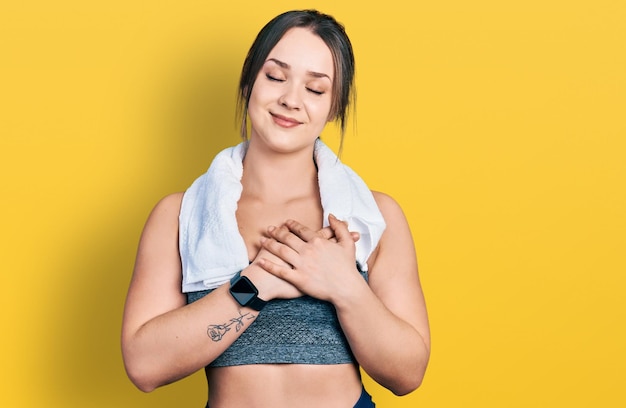 Young hispanic girl wearing sportswear and towel smiling with hands on chest with closed eyes and grateful gesture on face. health concept.