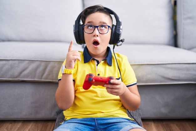 Giovane ragazzo ispanico che gioca al videogioco tenendo il controller con le cuffie stupito e sorpreso guardando in alto e indicando con le dita e le braccia alzate.