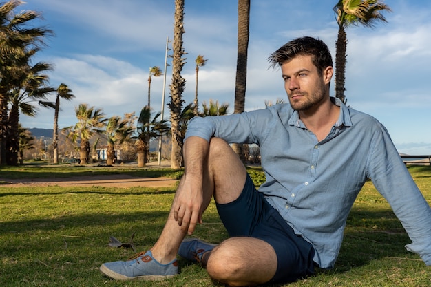 Free photo young hispanic male wearing a blue shirt sitting in a park with a lot of palm trees in background