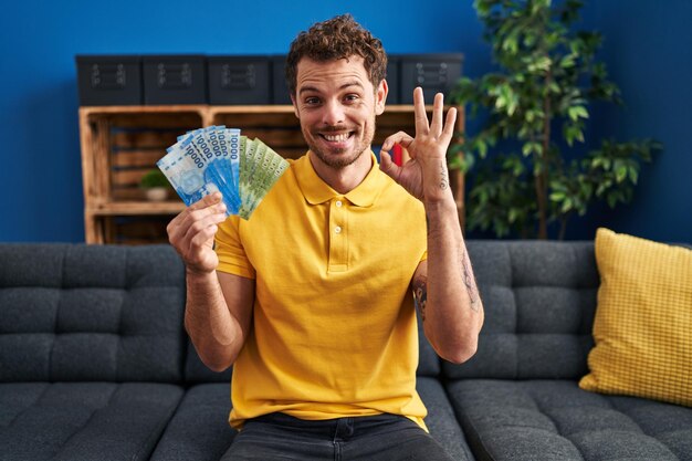 Young hispanic man holding chilean pesos doing ok sign with fingers smiling friendly gesturing excellent symbol