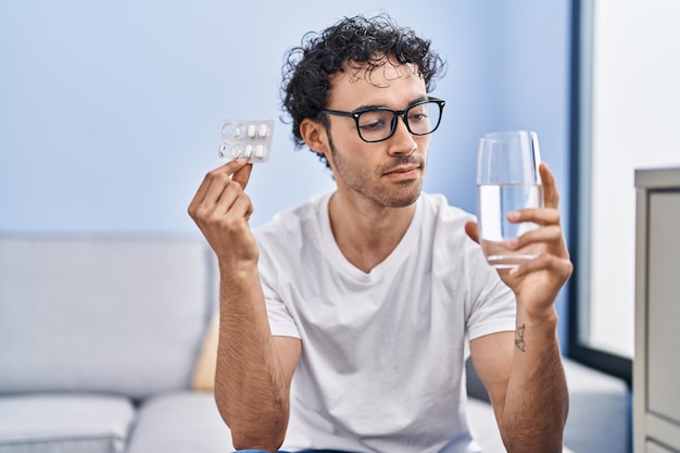 Foto gratuita giovane uomo ispanico che tiene pillole e bicchiere d'acqua a casa