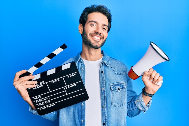 Foto gratuita giovane uomo ispanico che tiene l'assicella del film video e il megafono che sorride con un sorriso felice e fresco sul viso che mostra i denti