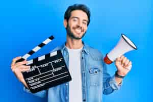 Foto gratuita giovane uomo ispanico che tiene l'assicella del film video e il megafono che sorride con un sorriso felice e fresco sul viso che mostra i denti