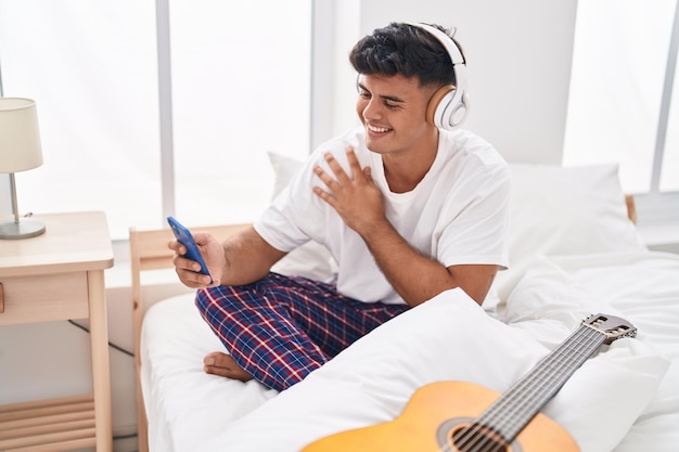 Young hispanic man listening to music sitting on bed at bedroom