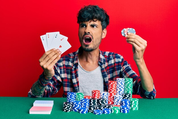 Young hispanic man playing poker holding cards and casino chips angry and mad screaming frustrated and furious, shouting with anger. rage and aggressive concept.