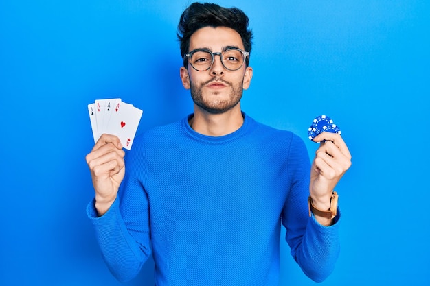 Young hispanic man playing poker holding casino chips and cards looking at the camera blowing a kiss being lovely and sexy love expression