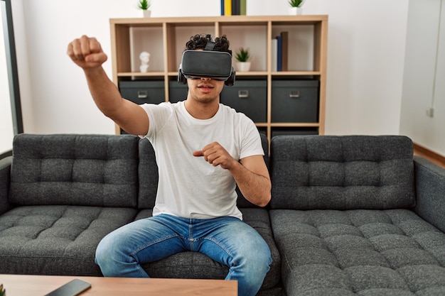 Young hispanic man playing with virtual reality glasses sitting on the sofa at home