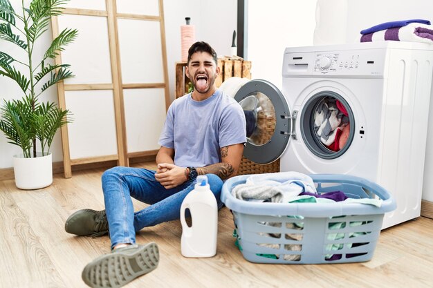 Young hispanic man putting dirty laundry into washing machine sticking tongue out happy with funny expression emotion concept