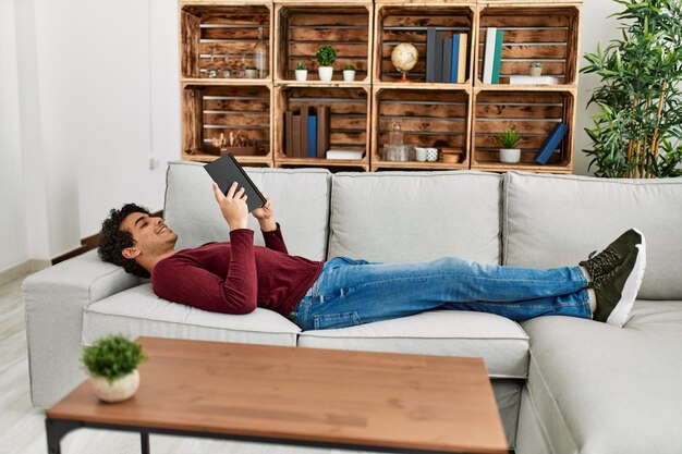 Молодой латиноамериканец читает книгу, лежа дома на диване