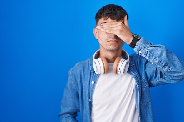 Giovane uomo ispanico in piedi su sfondo blu che copre gli occhi con la mano che sembra serio e triste concetto di nascondimento e rifiuto cieco