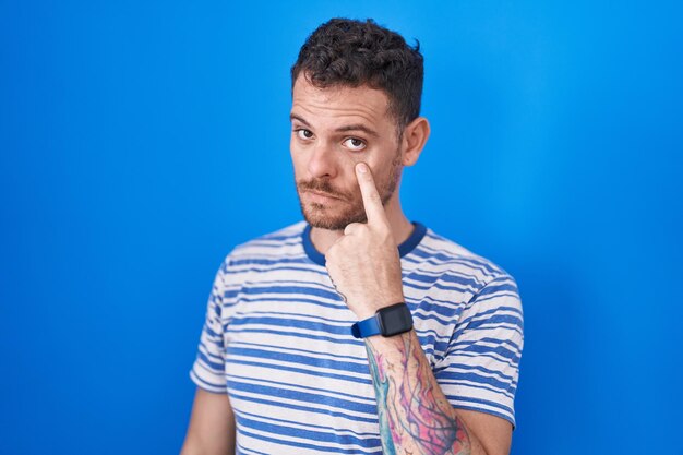 Young hispanic man standing over blue background pointing to the eye watching you gesture, suspicious expression