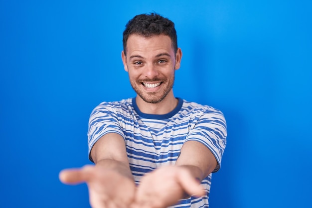 Young hispanic man standing over blue background smiling with hands palms together receiving or giving gesture hold and protection