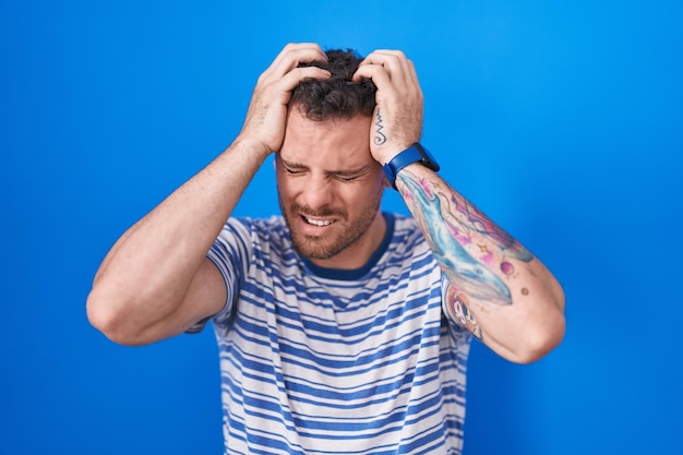 青い背景の上に立っている若いヒスパニック系の男性は、痛みと片頭痛のために必死でストレスを感じている頭痛に苦しんでいます。頭に手。