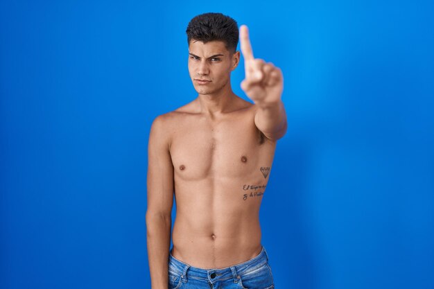 Young hispanic man standing shirtless over blue background pointing with finger up and angry expression showing no gesture
