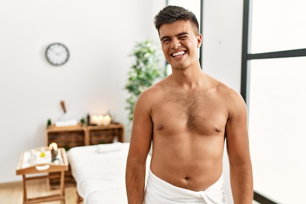 Free photo young hispanic man standing shirtless at spa center winking looking at the camera with sexy expression, cheerful and happy face.