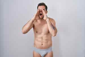 Free photo young hispanic man standing shirtless wearing underware shouting angry out loud with hands over mouth