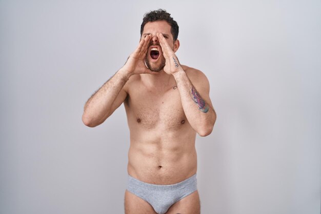 Young hispanic man standing shirtless wearing underware shouting angry out loud with hands over mouth