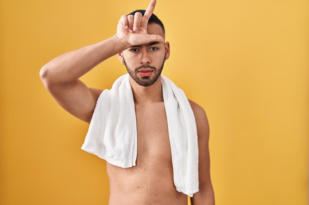 Молодой латиноамериканец, стоящий без рубашки с полотенцем, высмеивает людей с пальцами на лбу, делая жест неудачника, насмехаясь и оскорбляя.