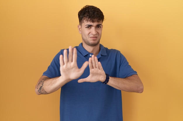 Young hispanic man standing over yellow background moving away hands palms showing refusal and denial with afraid and disgusting expression stop and forbidden