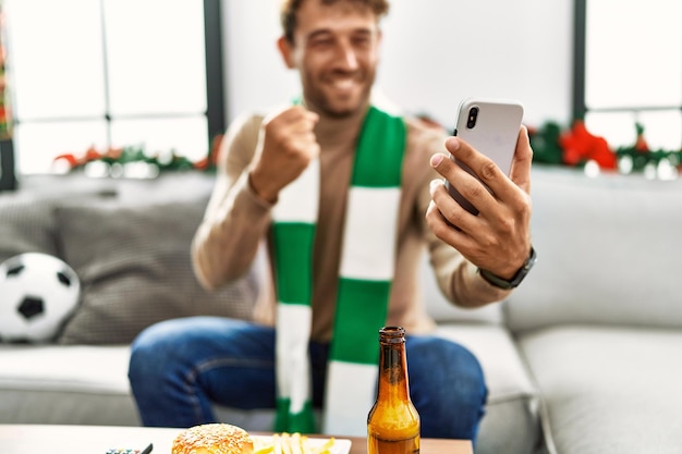 Foto gratuita giovane uomo ispanico che sostiene la partita di calcio utilizzando lo smartphone seduto vicino all'arredamento natalizio a casa