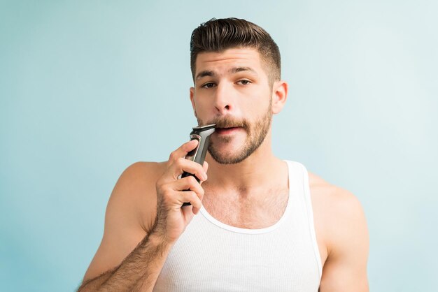 Young Hispanic man trimming mustache with electric razor while making eye contact against turquoise background