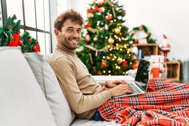 Free photo young hispanic man using laptop sitting by christmas tree at home