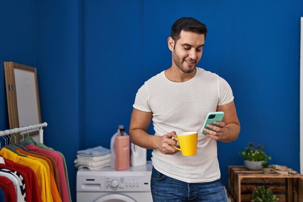 Young hispanic man using smartphone and drinking coffee waiting for washing machine at laundry room