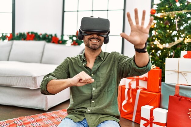 Young hispanic man using vr goggles sitting on floor by christmas tree at home