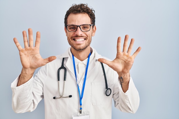 Молодой латиноамериканец в униформе врача и стетоскоп показывает и указывает пальцами номер десять, улыбаясь уверенно и счастливо.