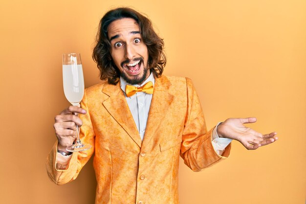 Молодой латиноамериканец в элегантном стиле ботаника пьет бокал игристого шампанского, празднуя достижение со счастливой улыбкой и выражением лица победителя с поднятой рукой