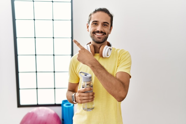 Young hispanic man wearing sportswear and drinking water at the gym cheerful with a smile on face pointing with hand and finger up to the side with happy and natural expression