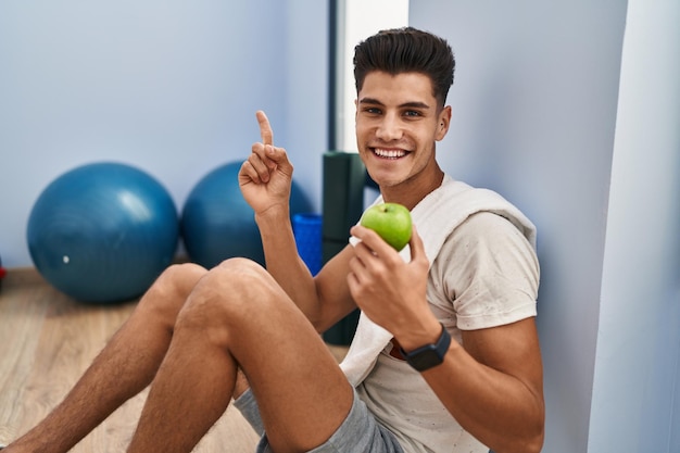 건강한 사과를 먹고 운동복을 입은 젊은 히스패닉 남자는 손과 손가락으로 옆을 가리키며 행복하게 웃고 있습니다.