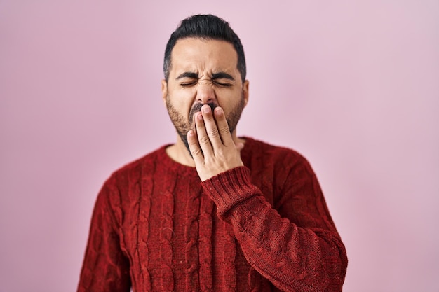 Бесплатное фото Молодой латиноамериканец с бородой в повседневном свитере на розовом фоне скучает, зевает, устал, прикрывая рот рукой, беспокойно и сонливо