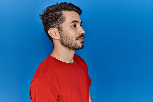 Молодой латиноамериканец с бородой в красной футболке на синем фоне смотрит в сторону, расслабляется в позе профиля с естественным лицом и уверенной улыбкой.