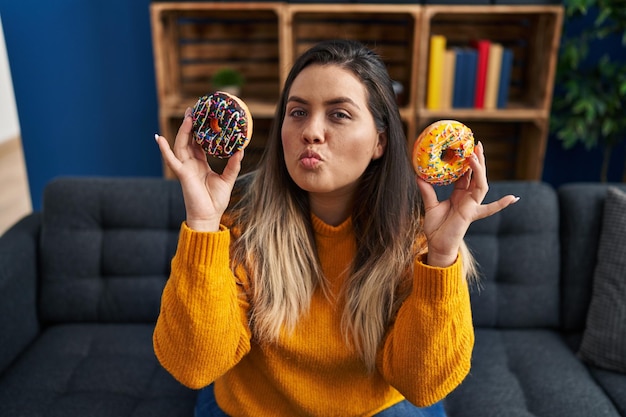 Free photo young hispanic woman eating doughnuts at home looking at the camera blowing a kiss being lovely and sexy love expression