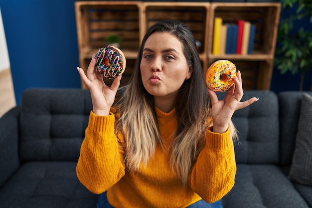 Foto gratuita giovane donna ispanica che mangia ciambelle a casa guardando la telecamera che soffia un bacio essendo espressione di amore adorabile e sexy