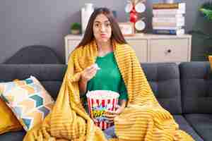 Free photo young hispanic woman eating snack sitting on the sofa at home puffing cheeks with funny face mouth inflated with air catching air