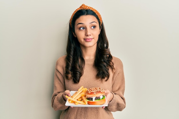 Giovane donna ispanica che mangia un gustoso hamburger classico sorridendo guardando di lato e fissando via il pensiero