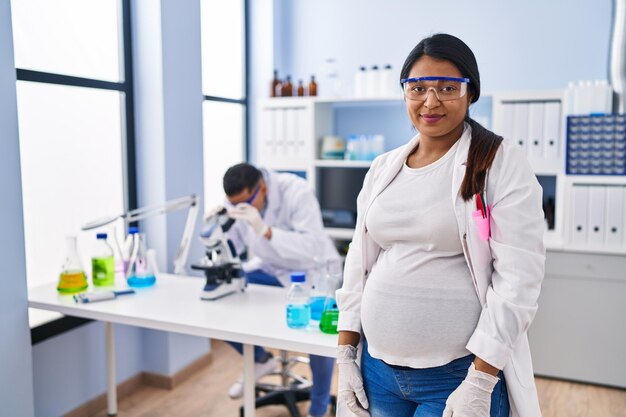 Молодая латиноамериканка, ожидающая ребенка, работает в научной лаборатории с серьезным выражением лица, просто и естественно смотрит в камеру