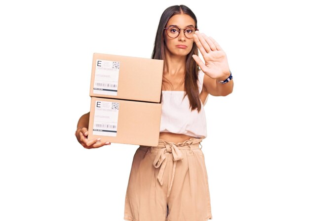 Young hispanic woman holding delivery package with open hand doing stop sign with serious and confident expression, defense gesture