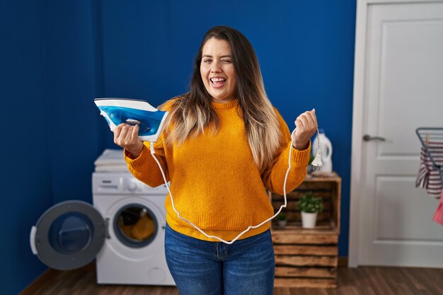 Young hispanic woman holding electric iron winking looking at the camera with sexy expression cheerful and happy face