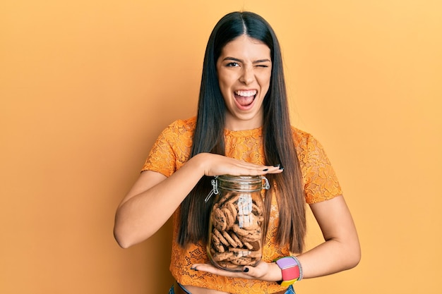 Free photo young hispanic woman holding jar with chocolate chips cookies winking looking at the camera with sexy expression cheerful and happy face